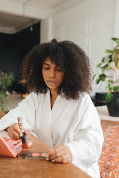 A woman with curly hair in a bathrobe applies makeup at a home setting.