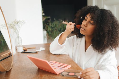 Free A woman with curly hair applies makeup at home using a brush and mirror, seated at a wooden table. Stock Photo