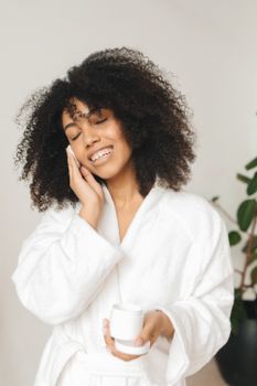 Smiling woman with curly hair applying face cream while wearing a bathrobe indoors.