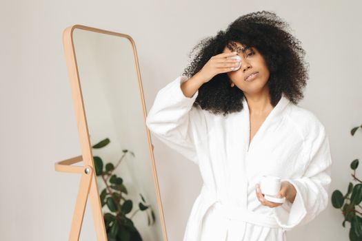 Woman in a white bathrobe using a cotton pad and holding a skincare container indoors.