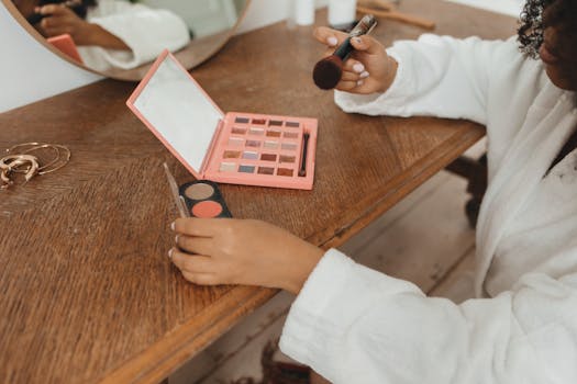 Woman wearing a bathrobe applying makeup at a home vanity table with brushes and cosmetics.
