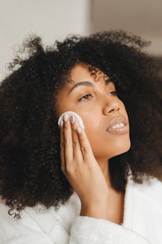 Side view of a woman cleansing her face with a cotton pad while wearing a bathrobe indoors.