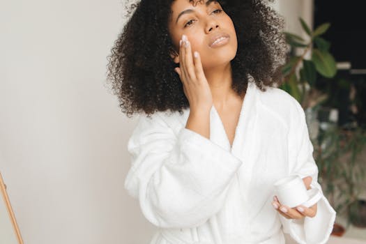 A woman with curly hair applies facial cream in a cozy home setting, promoting skincare routine.