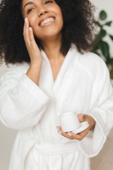 Smiling woman in bathrobe applying facial cream indoors, enjoying skincare routine.