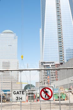 Construction site with no entry sign in front of towering modern skyscrapers under a clear blue sky.