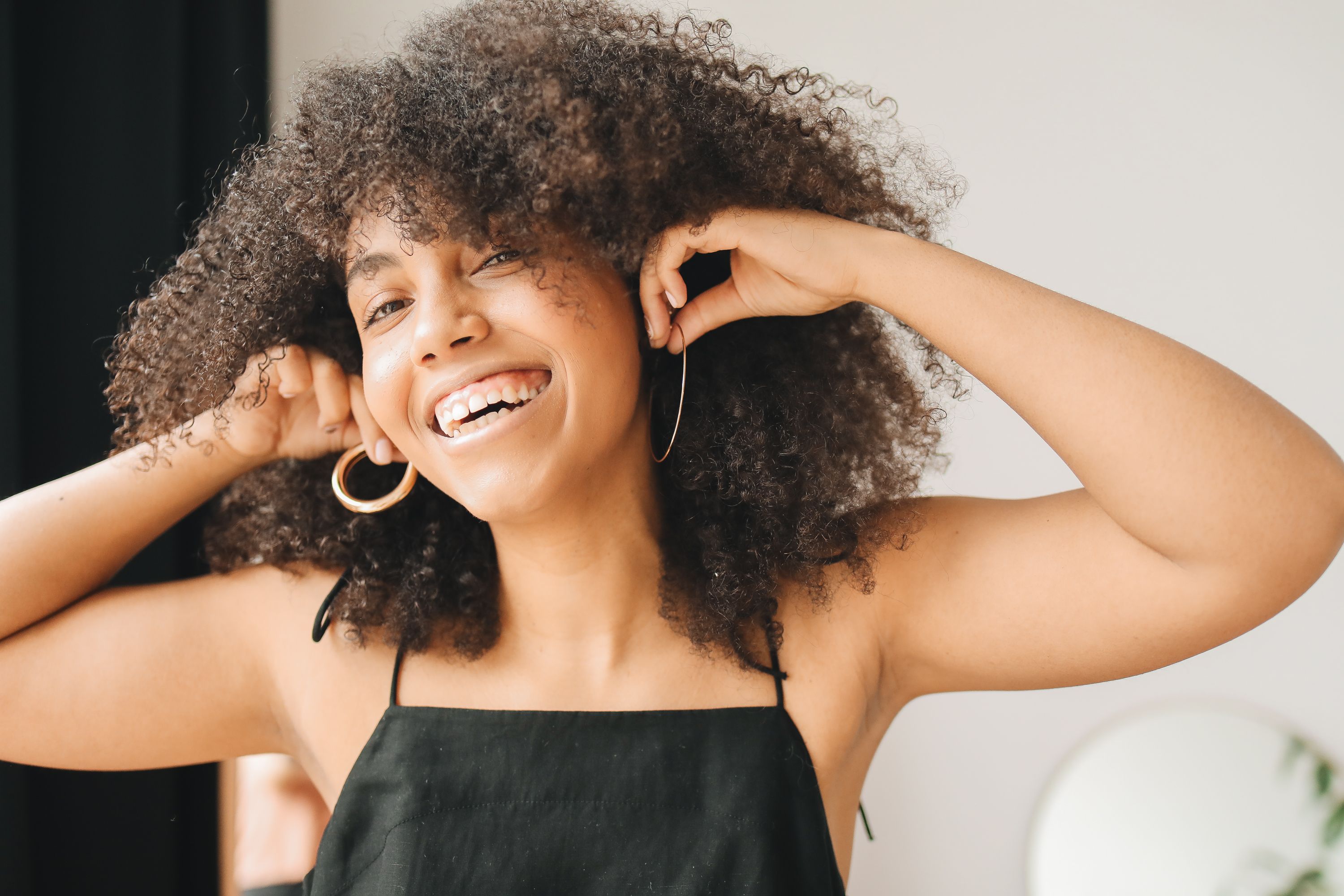 A happy woman with curly hair smiling while adjusting her earrings indoors.