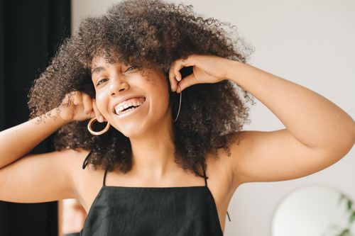 Free A happy woman with curly hair smiling while adjusting her earrings indoors. Stock Photo