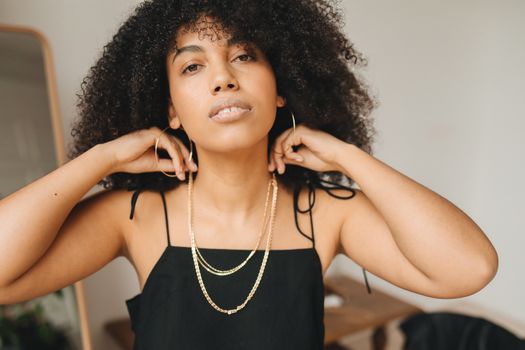 A woman in a black dress adjusting gold jewelry, embodying elegance and fashion indoors.