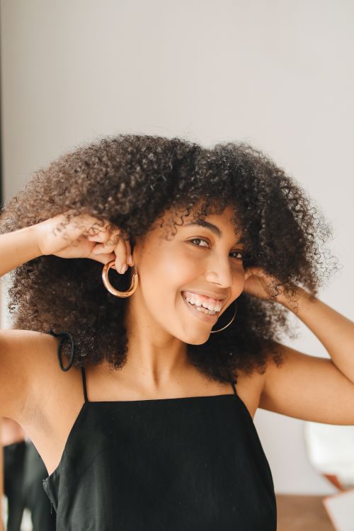 Free Confident woman in black dress adjusting earrings indoors with a joyful smile. Stock Photo