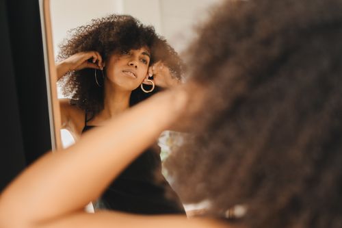 Free A woman in a black dress adjusts earrings while looking in the mirror, preparing for an event. Stock Photo