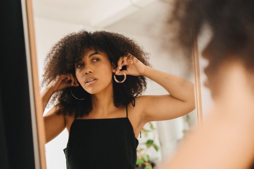 Free A woman adjusting her earrings while looking in the mirror, preparing for a social gathering. Stock Photo