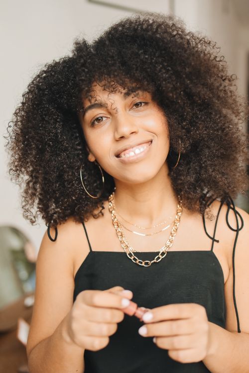 Free Stylish woman with curly hair smiling indoors wearing elegant jewelry and preparing to apply lipstick. Stock Photo