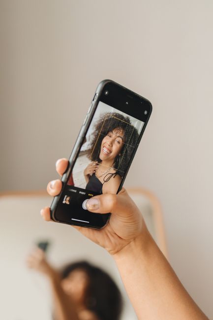 Black woman in elegant attire taking a selfie with a smartphone indoors.
