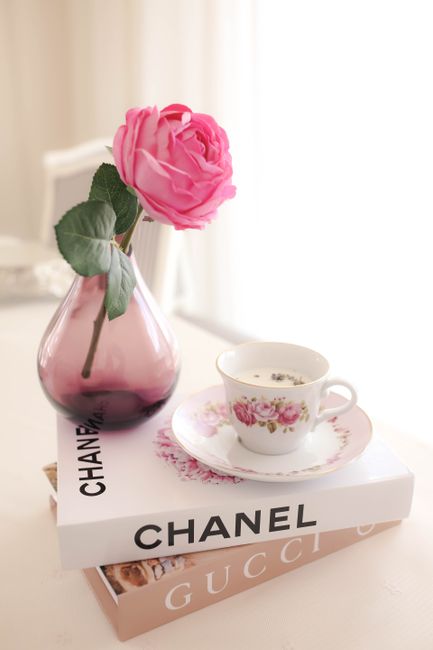 A delicate pink rose in a vase beside a floral tea cup on Chanel and Gucci books.
