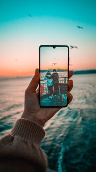 A hand holding a smartphone captures a couple by the ocean at sunset reflecting a serene moment.