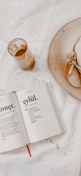 Stylish flat lay featuring an open book, glass of beverage, and sun hat on a soft fabric background.