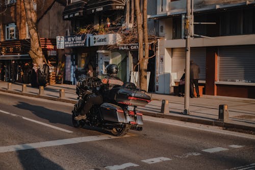 Back view of anonymous male biker in helmet riding modern motorbike on asphalt road along buildings on sunny street in city