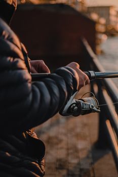 A man wearing a puffer jacket fishes at sunrise on a wooden pier, using a rod and reel.