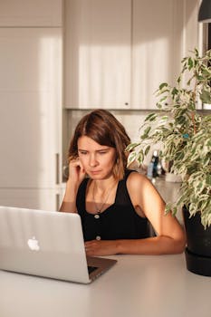 A woman in a black tank top works on her laptop in a bright, plant-filled kitchen, embodying remote work comfort.