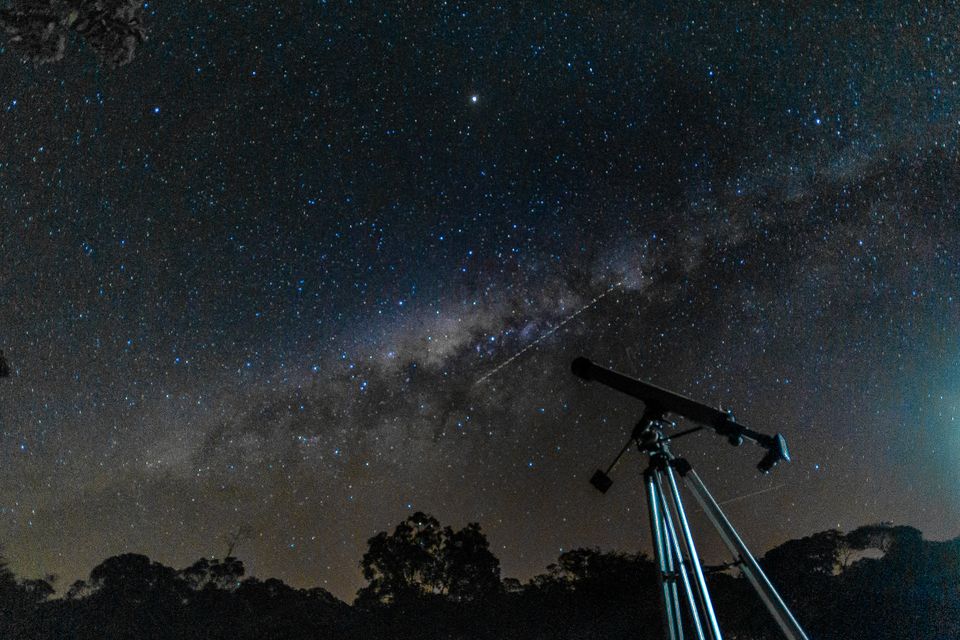 A telescope on a tripod in silhouette below a sky crossed by the Milky Way