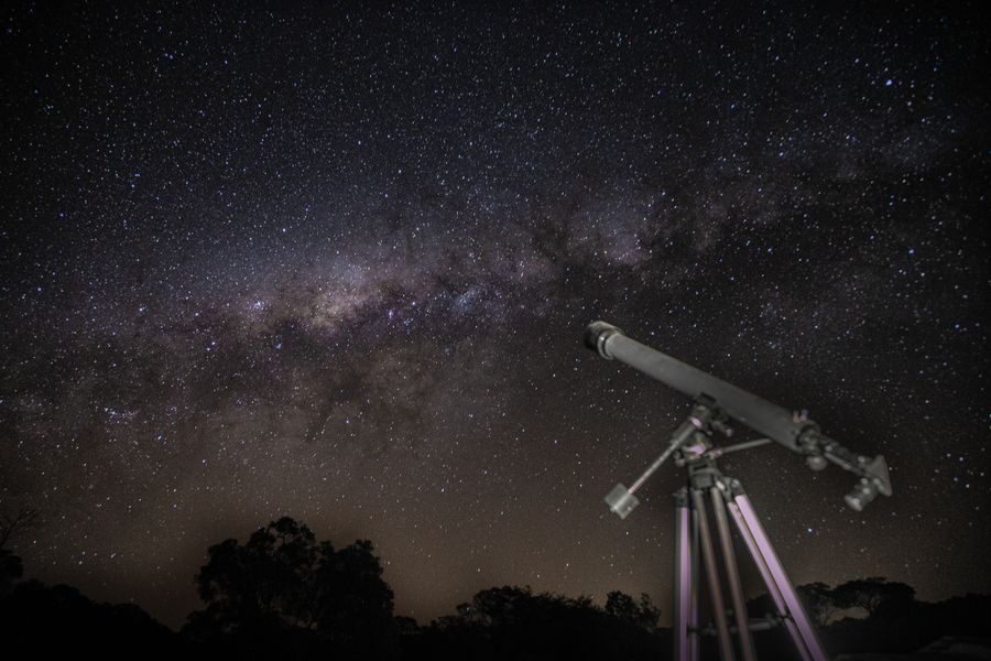 A small white refractor telescope on a tripod under a starry sky with trees on the horizon