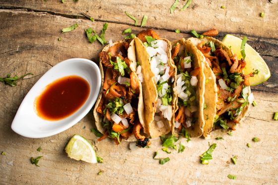 Four corn tacos with shredded red chicken, diced onion, cilantro, red salsa and lime wedges