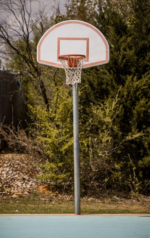 Free Basketball ring hanging on backboard against trees and bushes in park under clouds Stock Photo