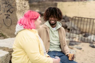 Cheerful multiracial couple with cans of soda sitting on stones on embankment near river while spending time together during date