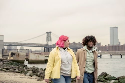 Free Multiethnic couple in outerwear strolling together on shore with stones near calm river against bridge and residential buildings in city Stock Photo