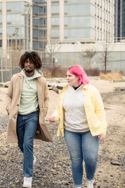 Free Multiracial couple in stylish outerwear walking on embankment with leafless trees against buildings during date in coastal area of city Stock Photo