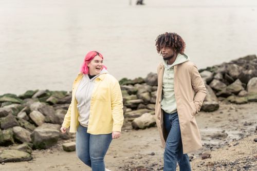 Free Positive plump woman with African American man in trendy outerwear strolling on waterside with stones near calm river during date Stock Photo