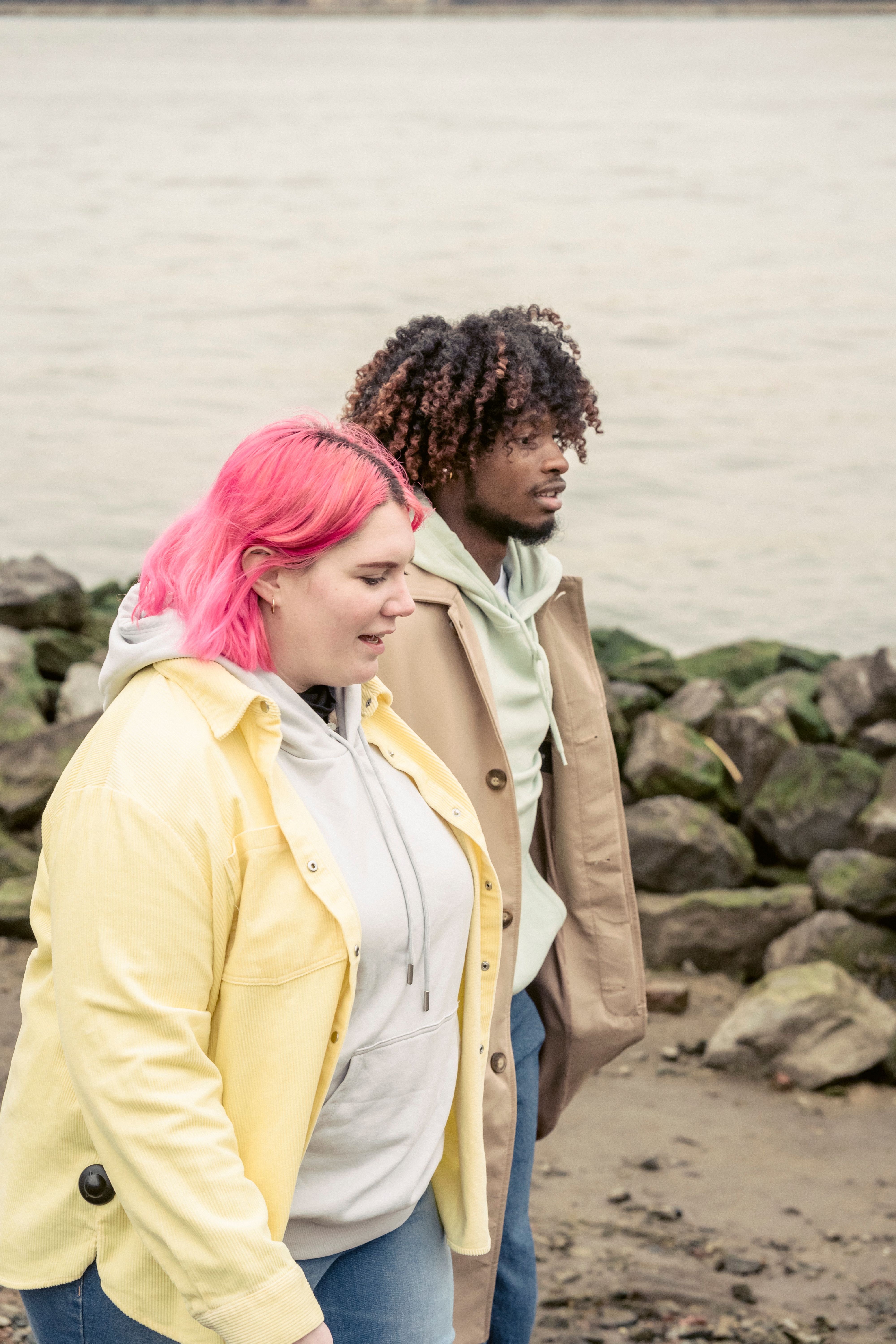 Side view of diverse couple in outerwear strolling on shore with stones near rippling water during date in coastal area