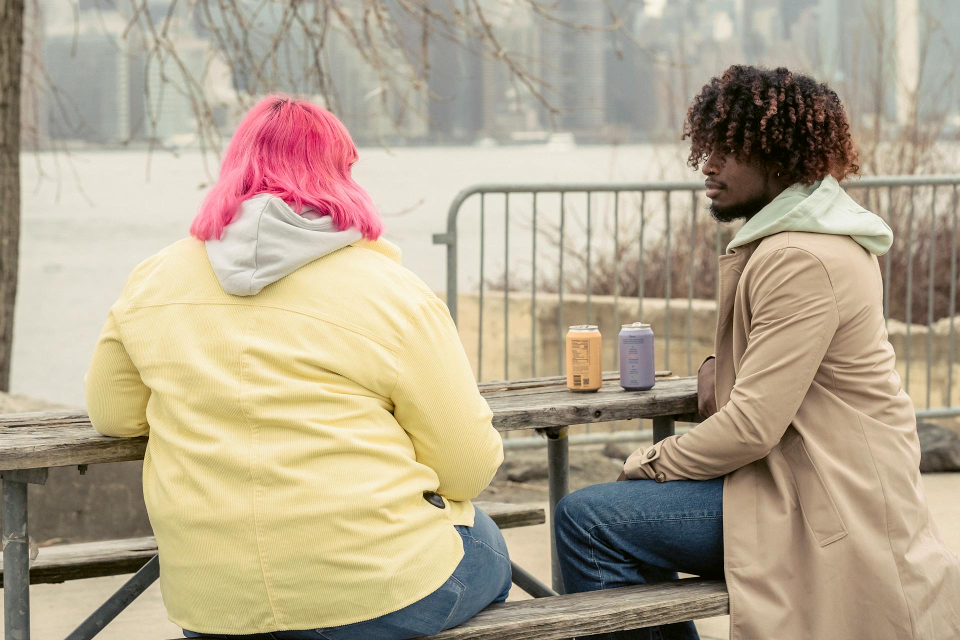 Stylish African American man and faceless woman with pink hair sitting at wooden table with cans of drinks while spending time on waterfront