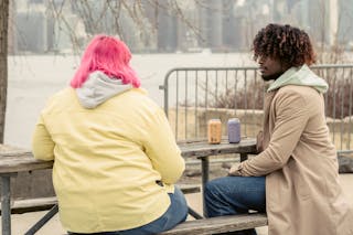 Stylish African American man and faceless woman with pink hair sitting at wooden table with cans of drinks while spending time on waterfront