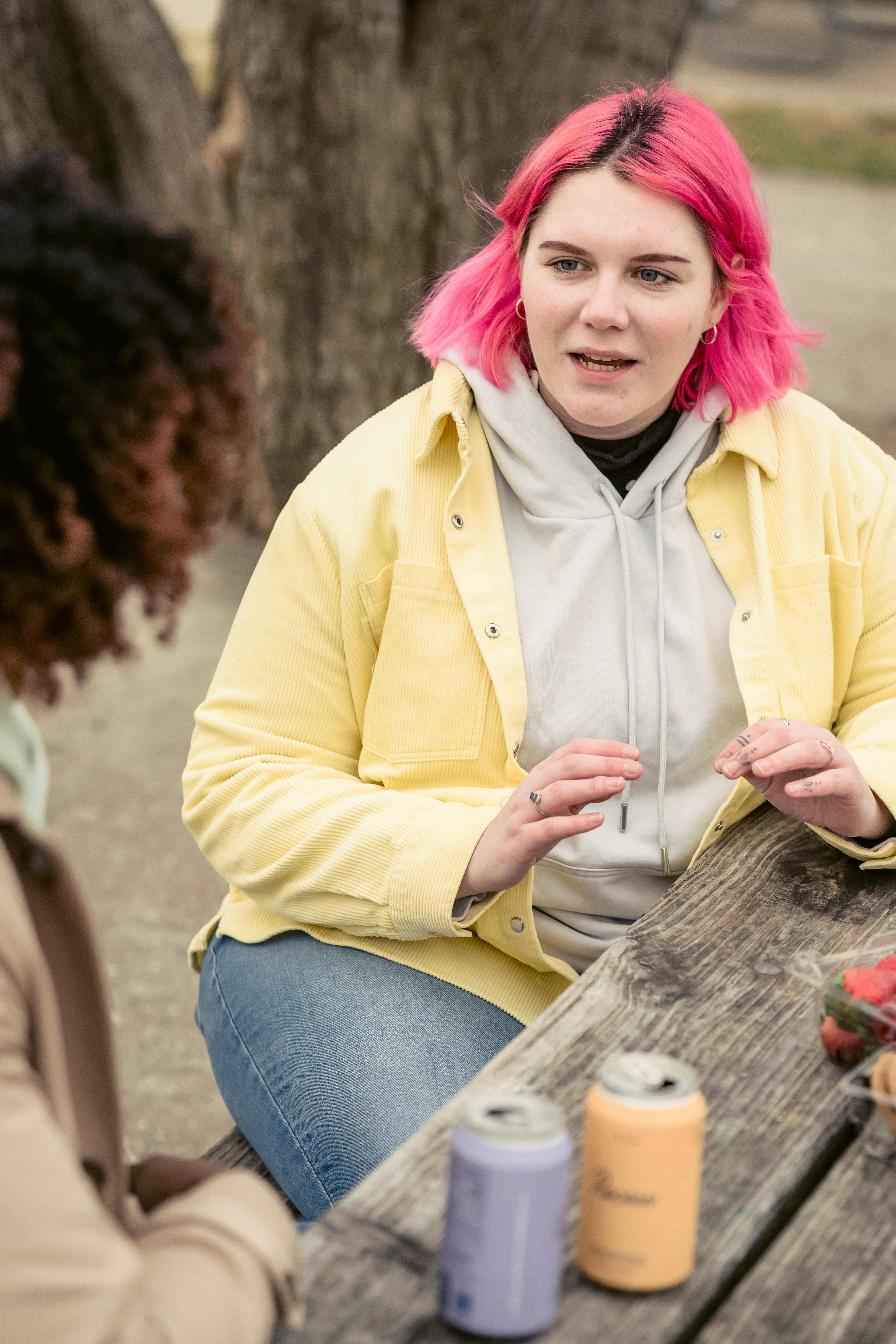 Woman with pink hair talking to crop anonymous man while sitting at wooden table with cans of soda on street