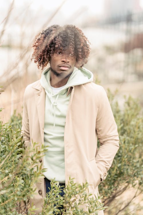 Serious African American male in stylish clothes looking away while standing on street with hand in pocket near lush shrubs on blurred background