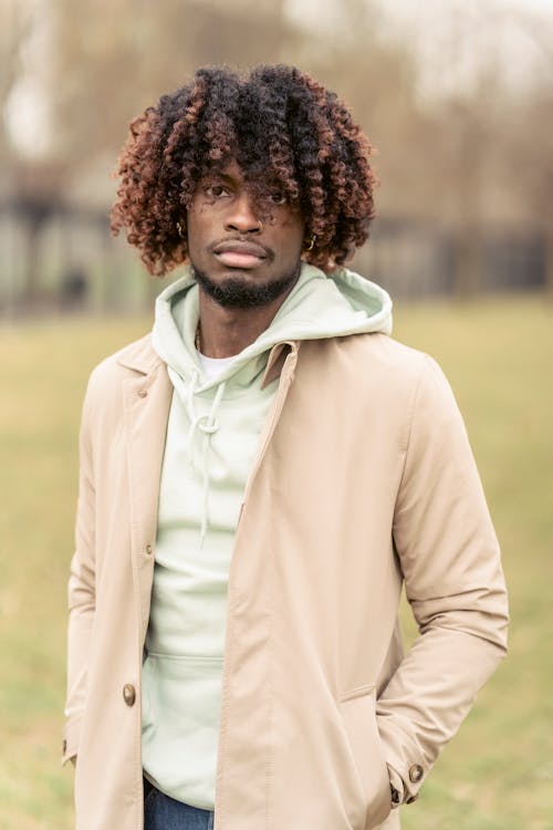 Serious African American male in trendy coat looking away while standing with hand in pockets on grassy ground in park on blurred background