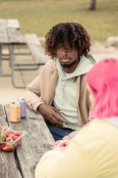 Calm African American man and faceless woman with pink hair sitting at wooden table with cans of soda and ripe strawberries on street