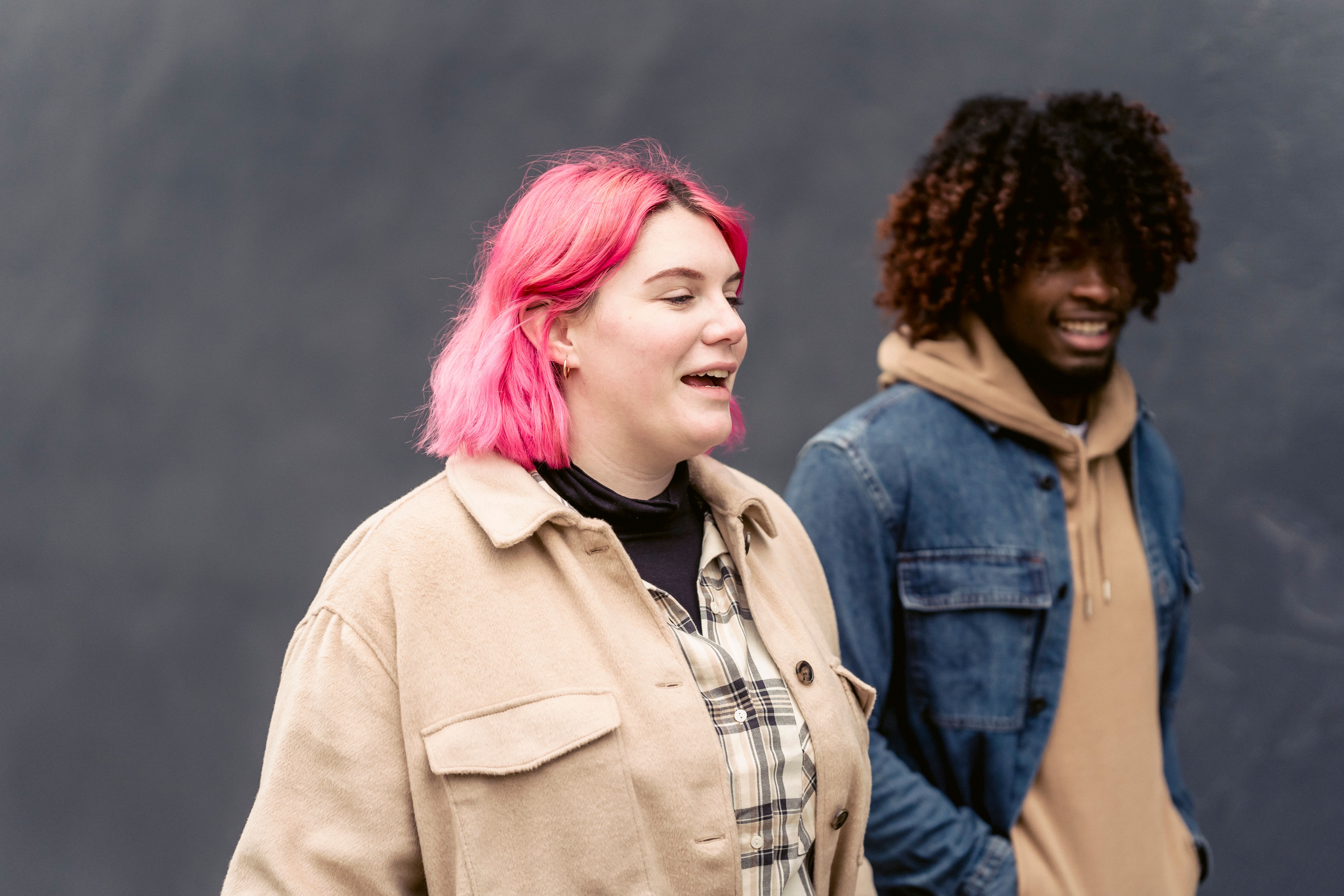 Free Cheerful woman with pink hair walking near African American man in trendy outfit along black wall on street during date Stock Photo
