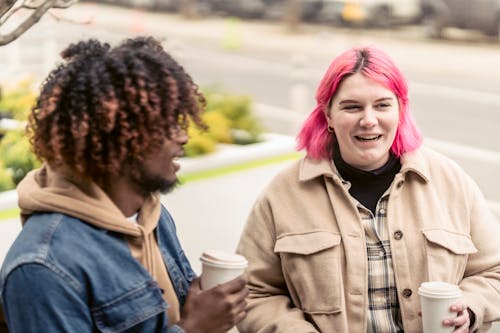Cheerful young female teenager with dyed pink hair in stylish warm clothes smiling and drinking coffee to go while speaking with positive black male friend in park