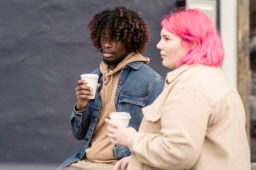 Trendy young African American guy with curly hair in stylish outfit drinking takeaway coffee and speaking with calm female friend while sitting together on bench on street