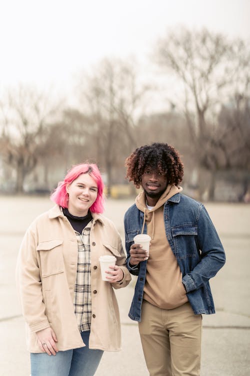 Free Positive young woman with pink hair and African American man smiling and looking at camera while standing on city square and drinking takeaway coffee Stock Photo