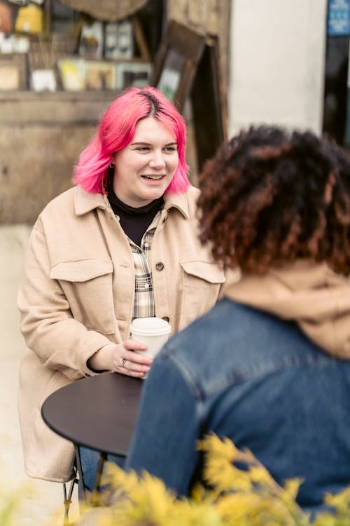 Happy female with pink hair smiling and speaking with man while sitting at table and enjoying coffee to go in street cafe