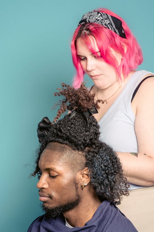Free Serious female hairdresser dying hair of African American male sitting in cape against blue background Stock Photo