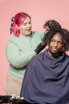 Female hairdresser with bright dyed hair dying hair of African American male customer sitting against pink wall