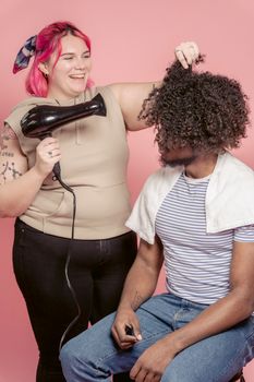 Laughing female hairstylist with hair dryer doing hairstyle to African American curly haired customer in studio against pink background