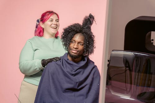 Free Positive African American female hairdresser hairstyling African American male customer in studio with light pink wall Stock Photo