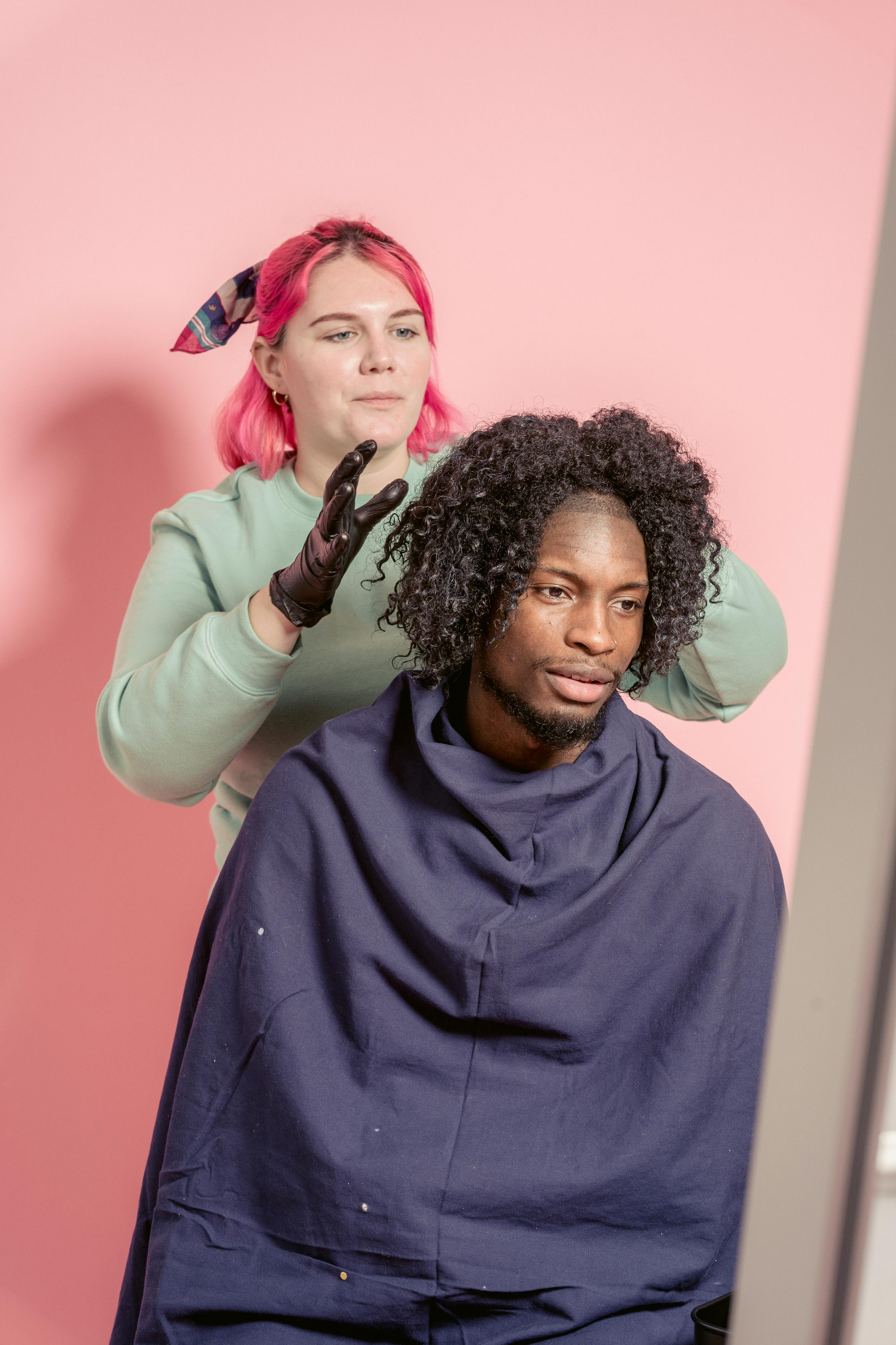 Female hairstylist standing behind bearded African American male client wrapped in cape while touching hair