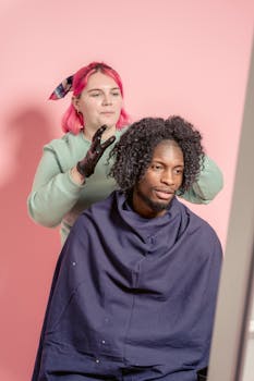 Female hairstylist standing behind bearded African American male client wrapped in cape while touching hair