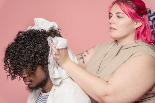 Serious female hairdresser wiping hair of African American client with towel while standing on pink background in modern light studio